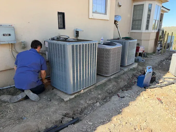 Technician kneeling beside outdoor HVAC units along a house foundation, working on a service trench
