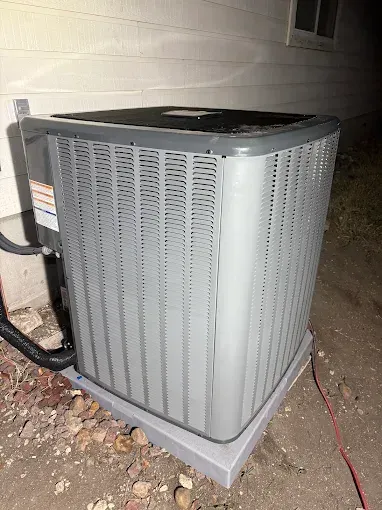 Outdoor air conditioner unit beside a house at night, with a red cord on the ground.