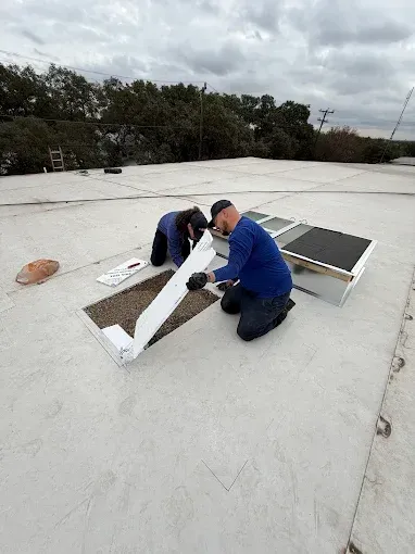 Two workers install a rooftop skylight on a flat white roof under cloudy skies.