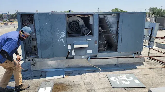 Worker inspecting a large blue HVAC unit outdoors on a rooftop.