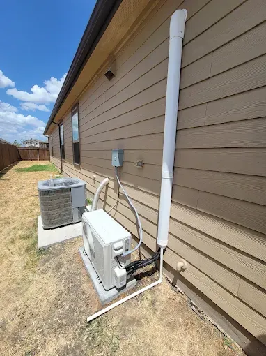Outdoor home HVAC unit beside tan siding with a white vertical vent pipe and air conditioner under a blue sky