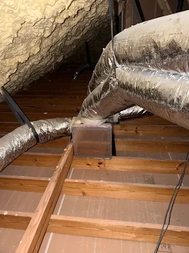 Insulated silver ductwork in an attic with exposed wooden rafters and floor joists.