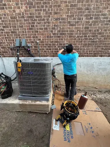 Worker in blue shirt taking a photo of HVAC unit beside a brick wall.