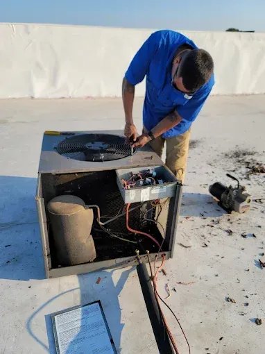 Technician in blue shirt repairing an outdoor air-conditioning unit on a rooftop.