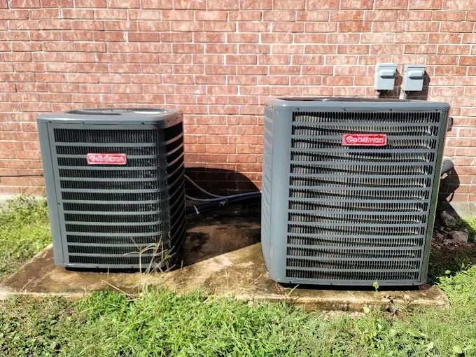 Two outdoor air conditioner units beside a brick wall on a concrete pad