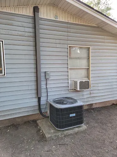 Side of a gray house with a window, wall vent, and a central air conditioner on gravel.