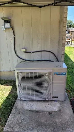 Outdoor window AC unit on a concrete pad beside a beige wall, with hose and electrical cable attached.