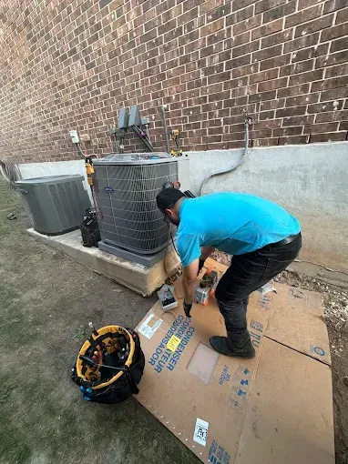 Person kneeling beside HVAC units outdoors, taping cardboard on the ground near a brick wall.