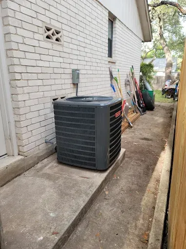 Outdoor HVAC unit beside a brick house, with tools and boards leaning against the wall along a narrow side yard