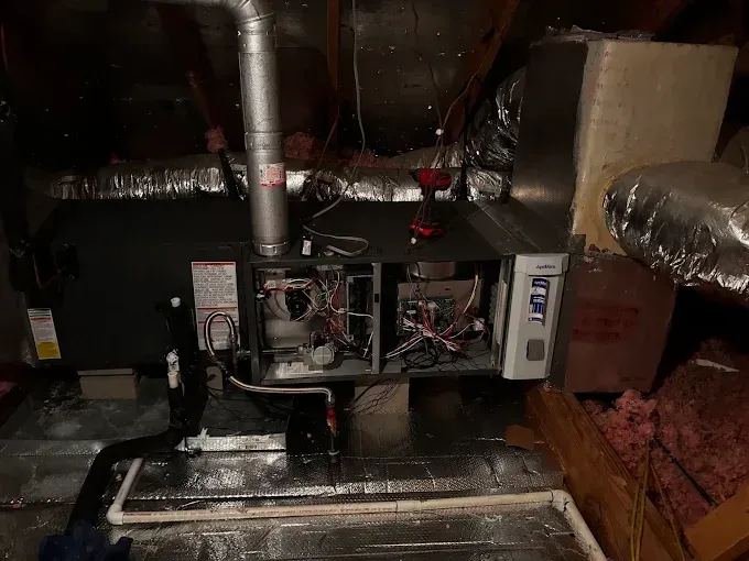 HVAC furnace and ductwork in a dim attic, with exposed wiring and metal vents.