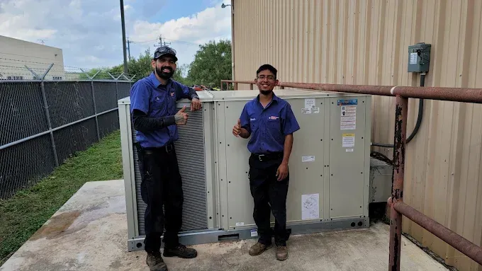 Two technicians in blue shirts stand beside an outdoor HVAC unit next to a beige building.