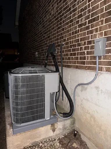 Outdoor HVAC condenser unit beside a brick wall with exposed wiring and conduit