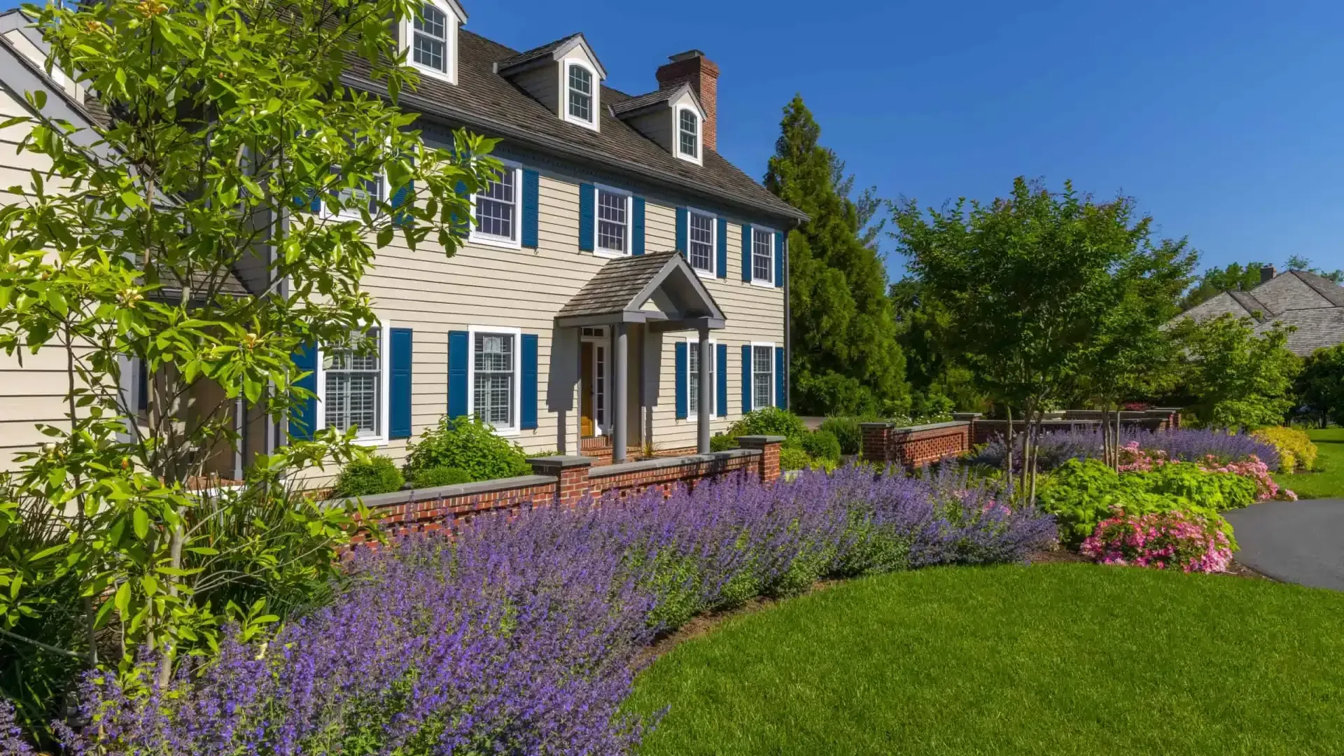 Suburban house with blue shutters, front porch, and a colorful flower garden under a clear blue sky