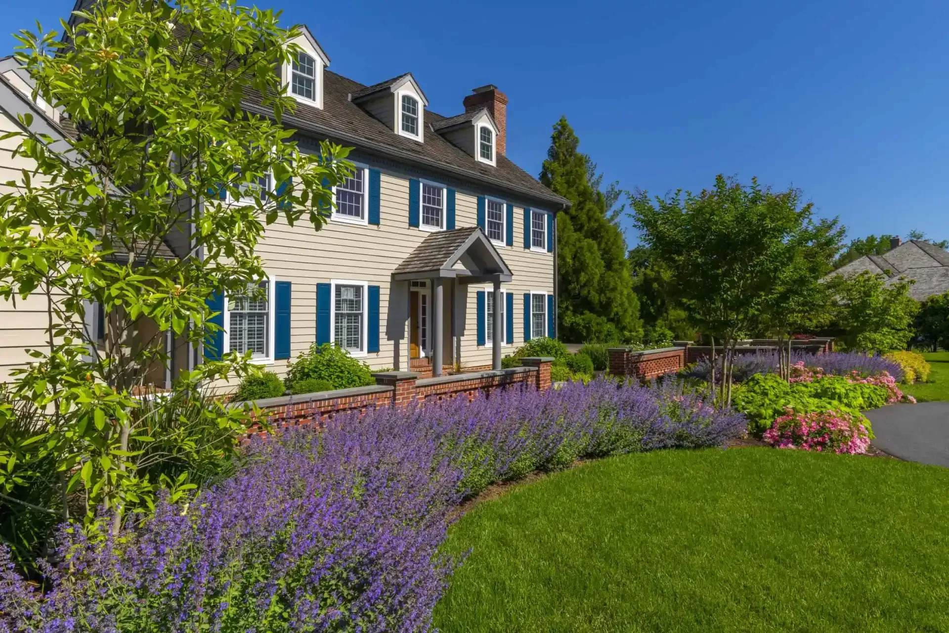 Large white house with blue shutters, a manicured lawn, and purple flower beds under a clear blue sky