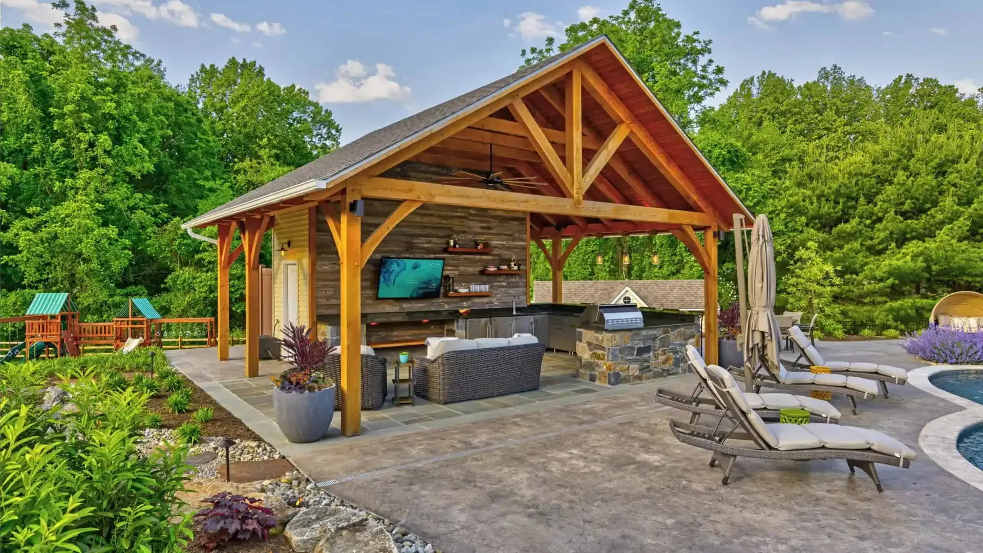 Outdoor patio with a wooden pavilion, stone fireplace, grill, and lounge chairs beside a pool and trees
