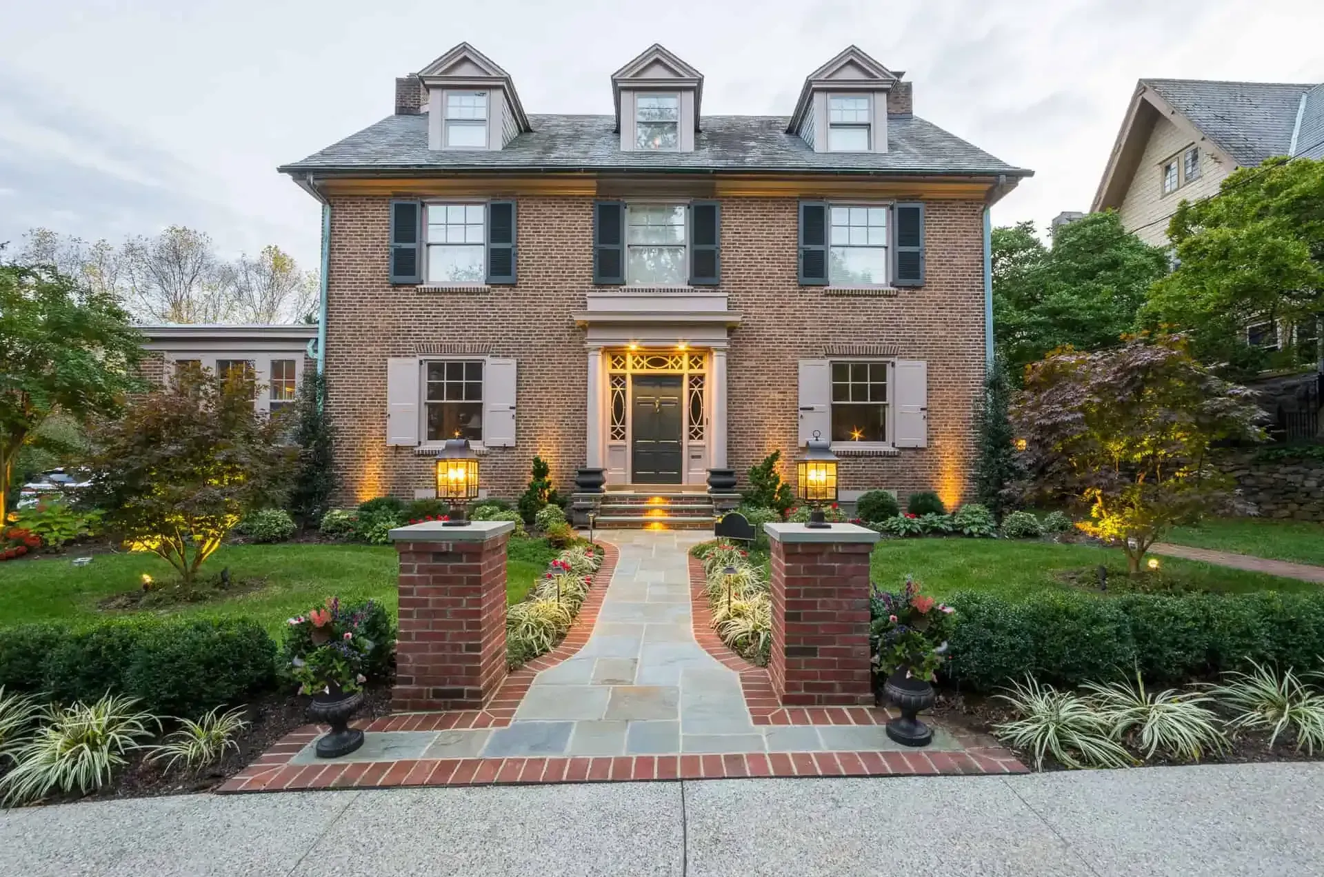 Brick two-story house with lit front entry, dormer windows, and landscaped walkway at dusk