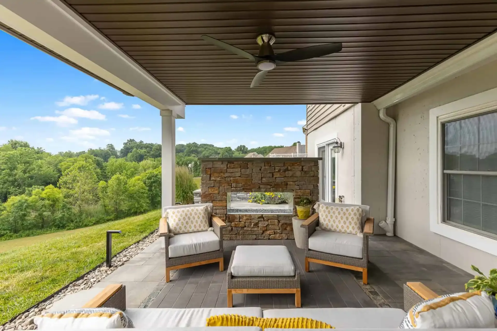 Covered patio with wicker seating, ottoman, ceiling fan, and a view of green fields.