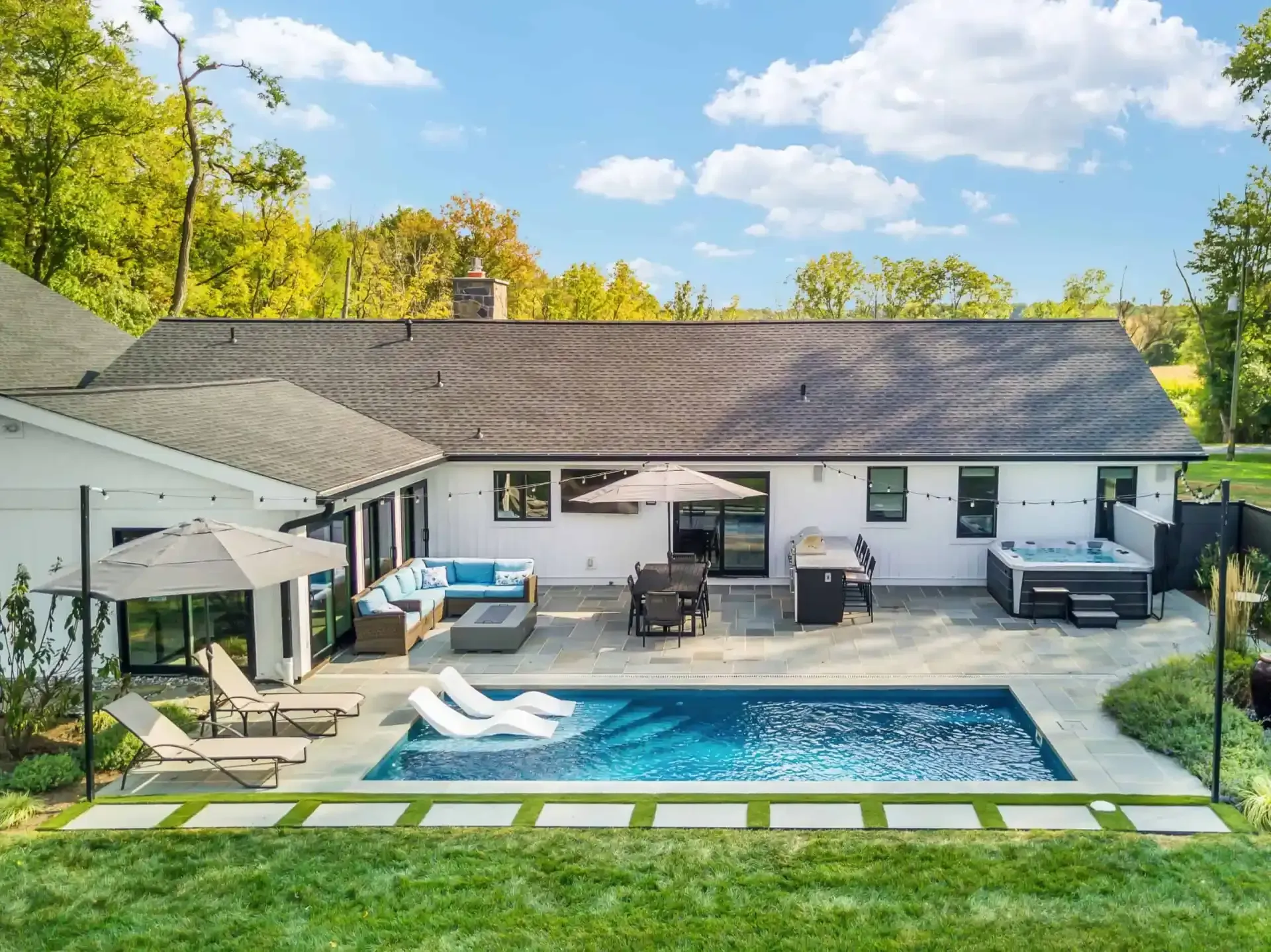 Backyard with a modern house, blue pool, patio seating, and green lawn under a bright sky