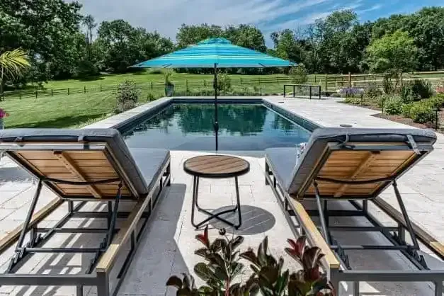 Modern patio with two lounge chairs, round table, and turquoise umbrella beside a pool and green lawn