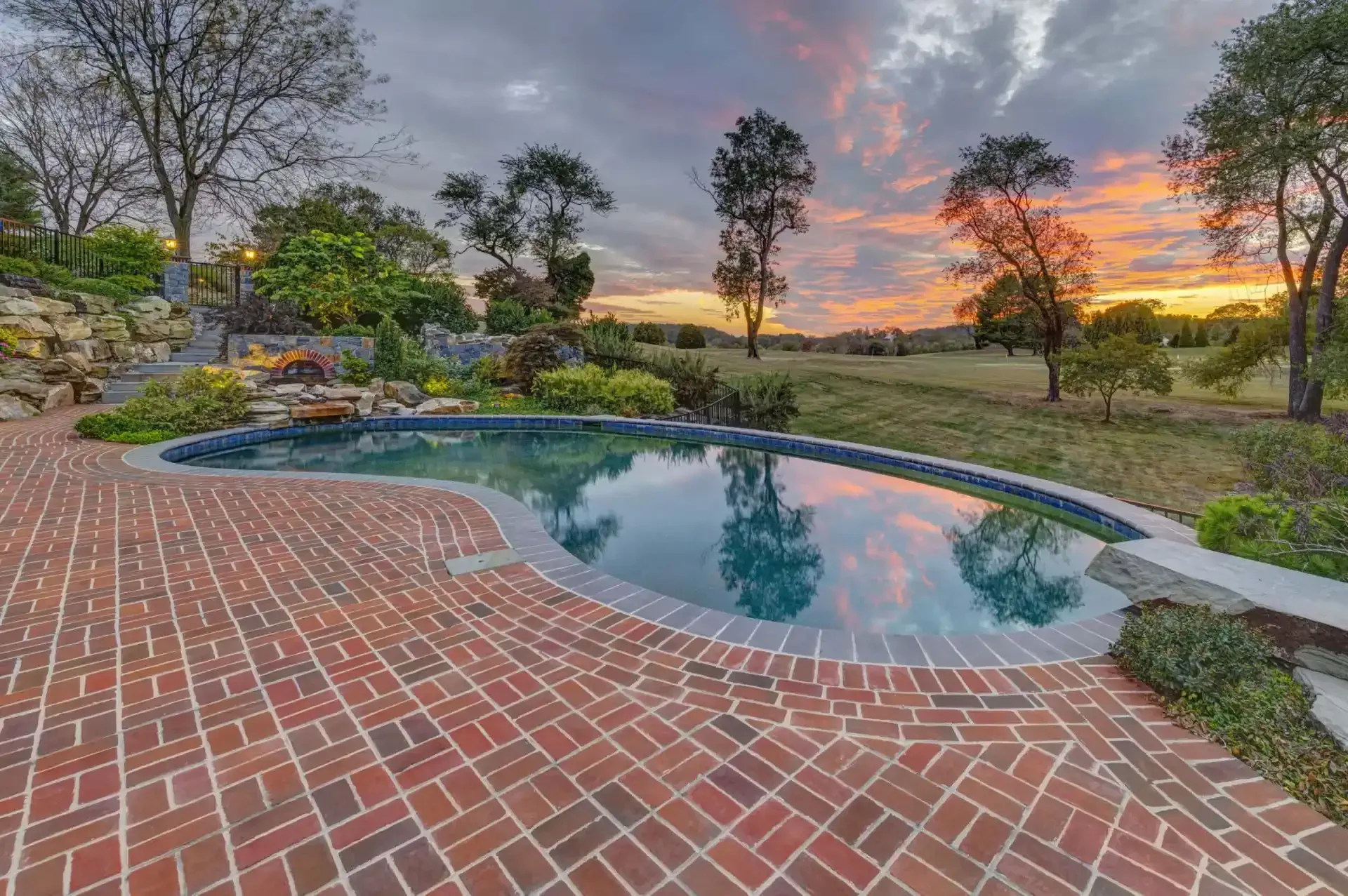 Brick patio beside a small backyard pool at sunset, with trees and a colorful sky.