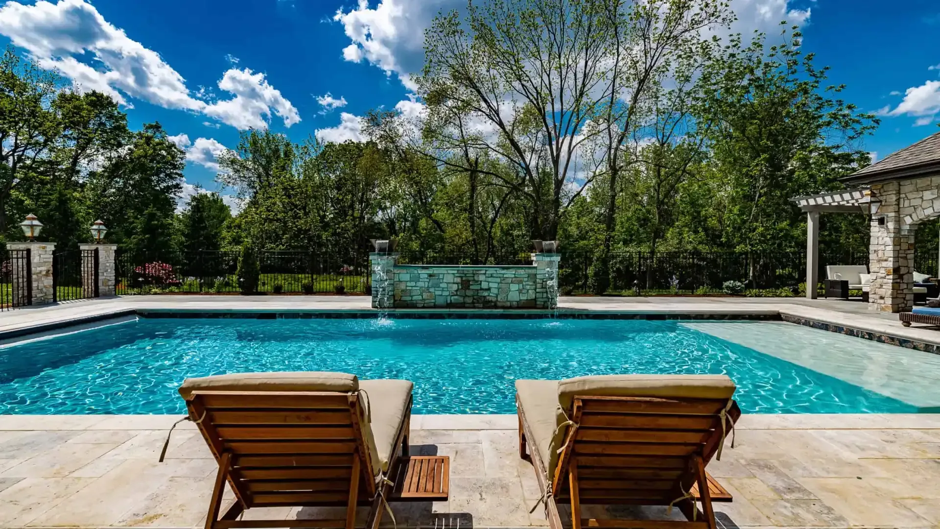 Backyard pool with two wooden lounge chairs, stone patio, trees, and bright blue sky