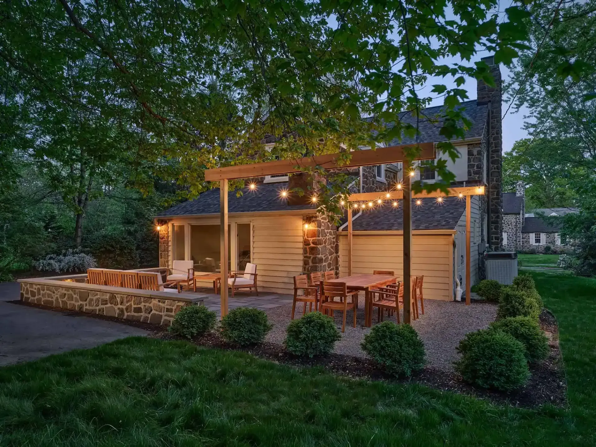 Cozy backyard patio with string lights, wooden pergola, seating, and stone retaining wall at dusk