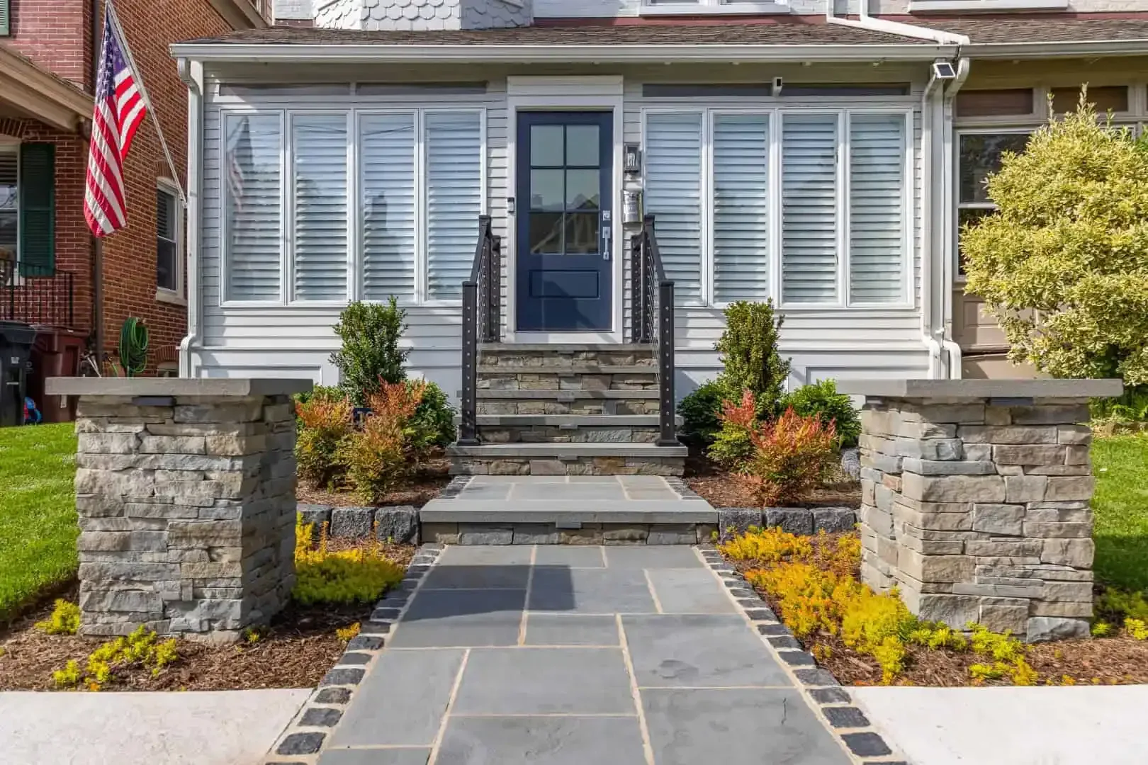 Front view of a stone walkway leading to a white house with steps, shrubs, and a U.S. flag.