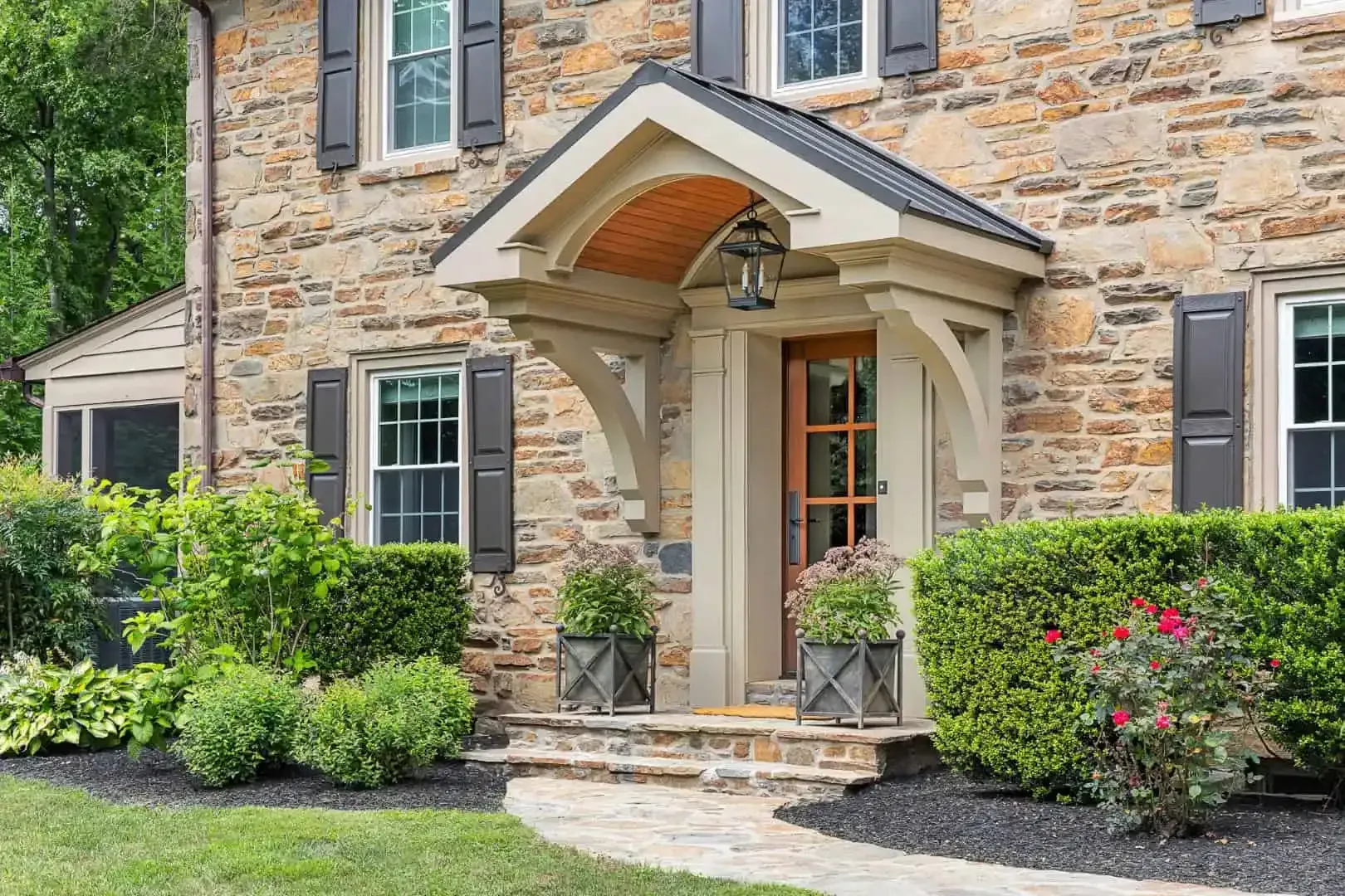 Stone house front entrance with arched porch, black shutters, and landscaped garden