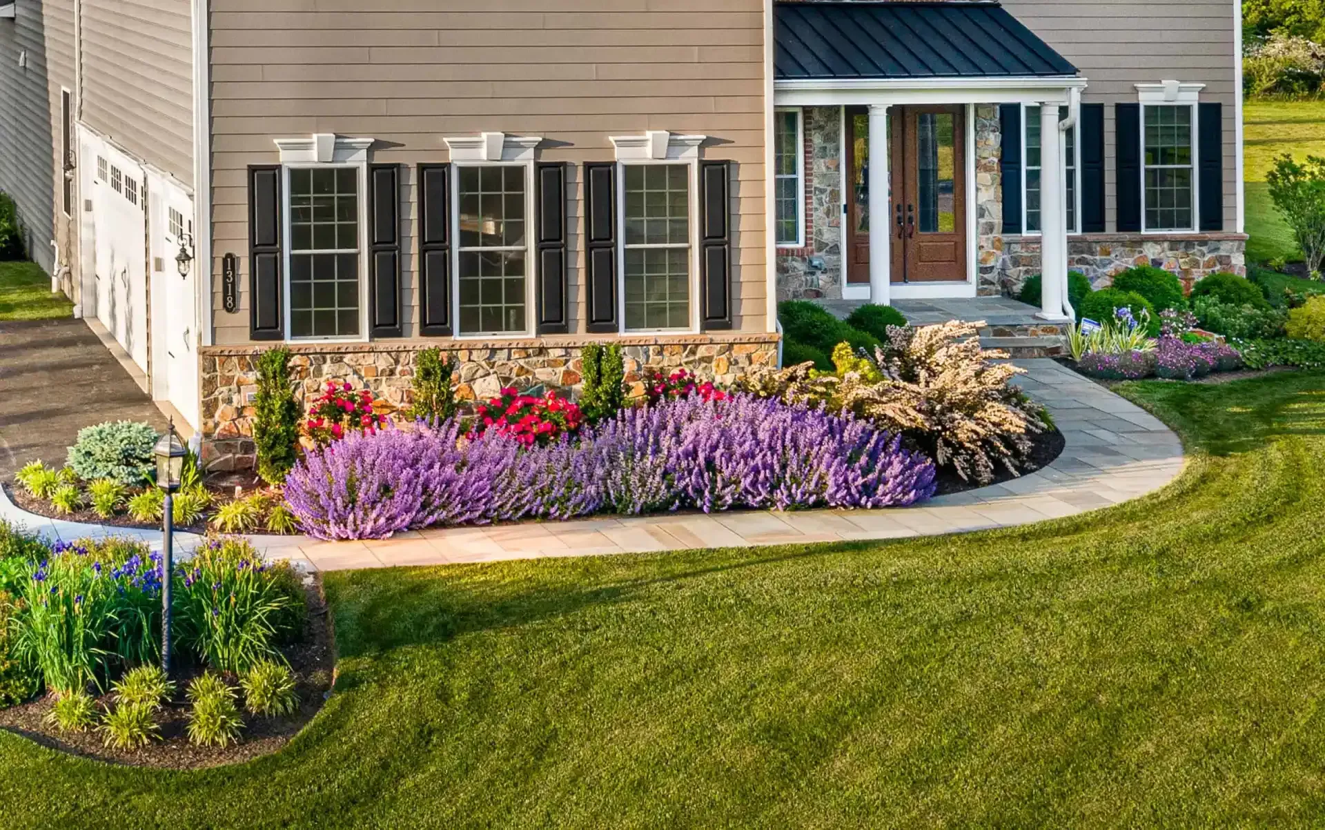 Suburban house with a curved flower bed of purple, pink, and yellow blooms beside a green lawn.
