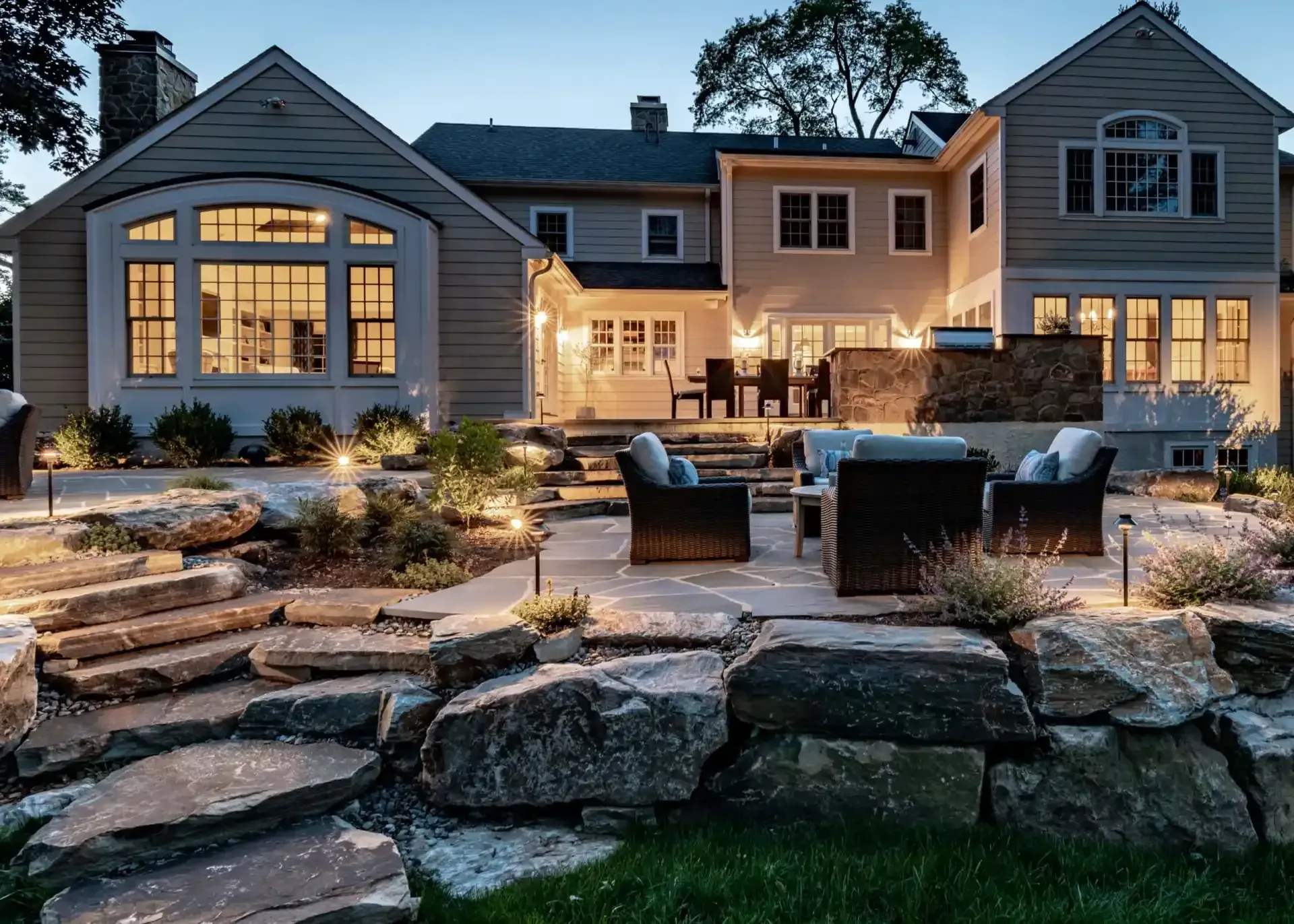 Luxury house at dusk with lit windows, stone patio seating, and landscaped backyard steps leading to the lawn