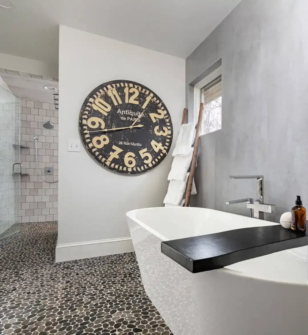 Modern bathroom with freestanding tub, large wall clock, pebble floor, and gray walls