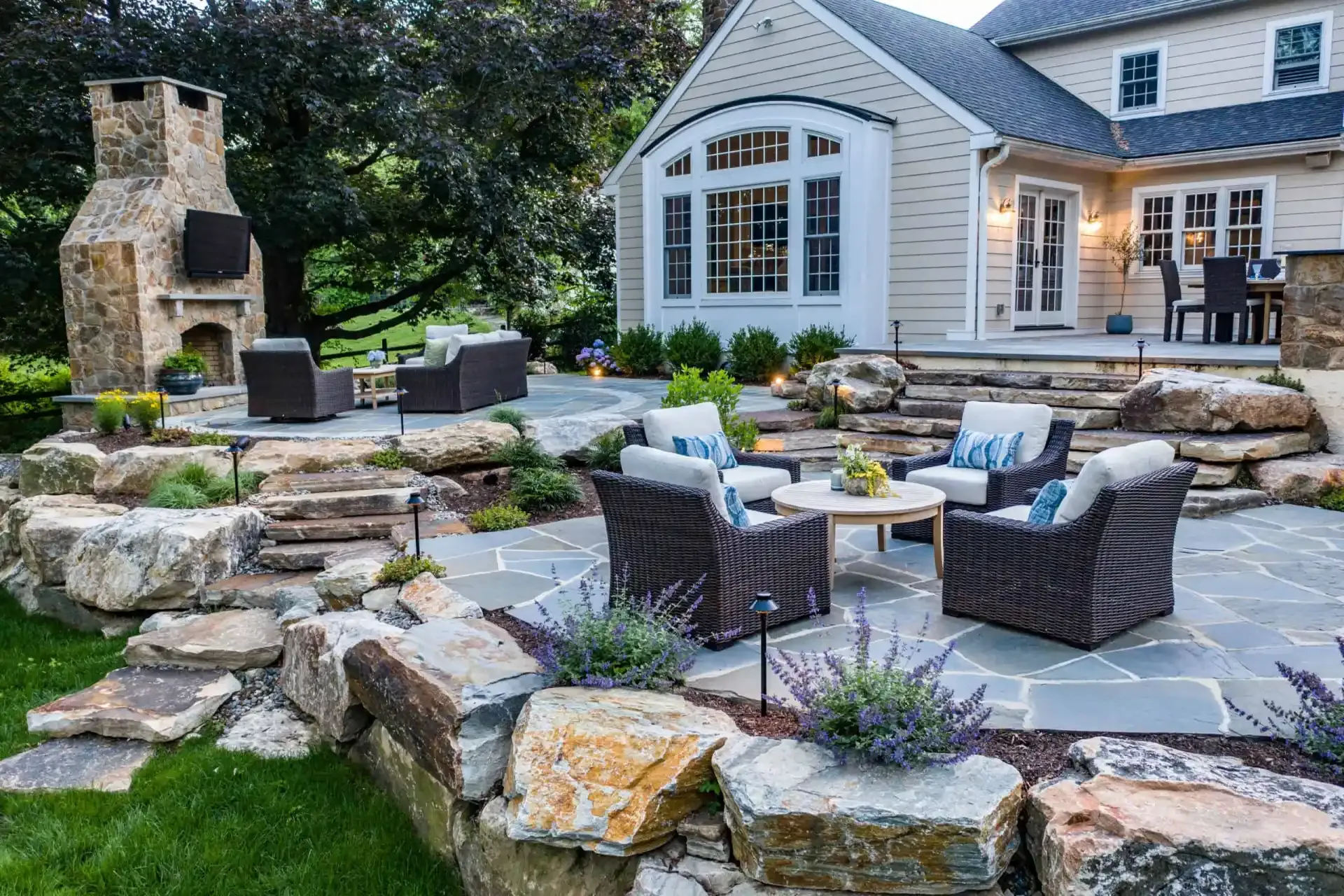 Stone patio with wicker chairs and fire pit beside a white house and outdoor fireplace