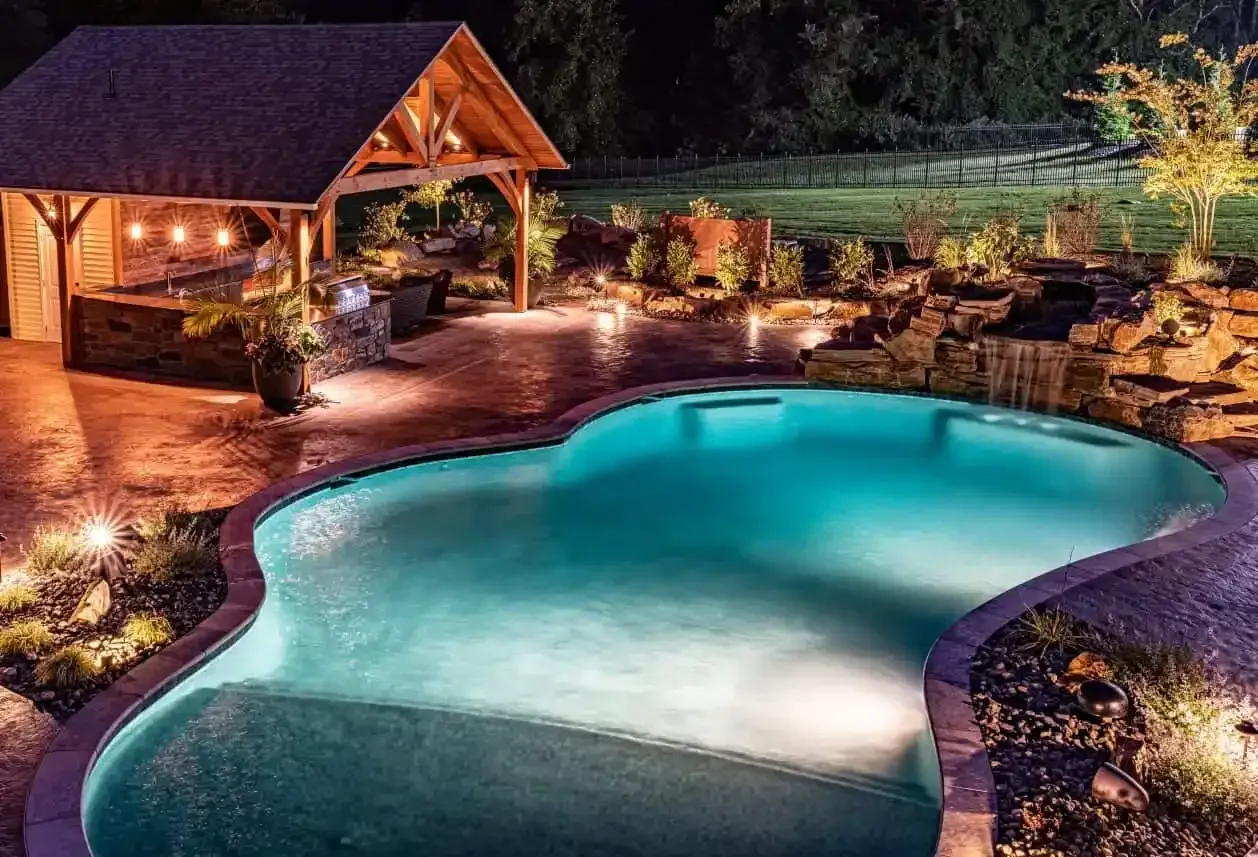 Illuminated backyard pool with a wooden gazebo and stone patio at night