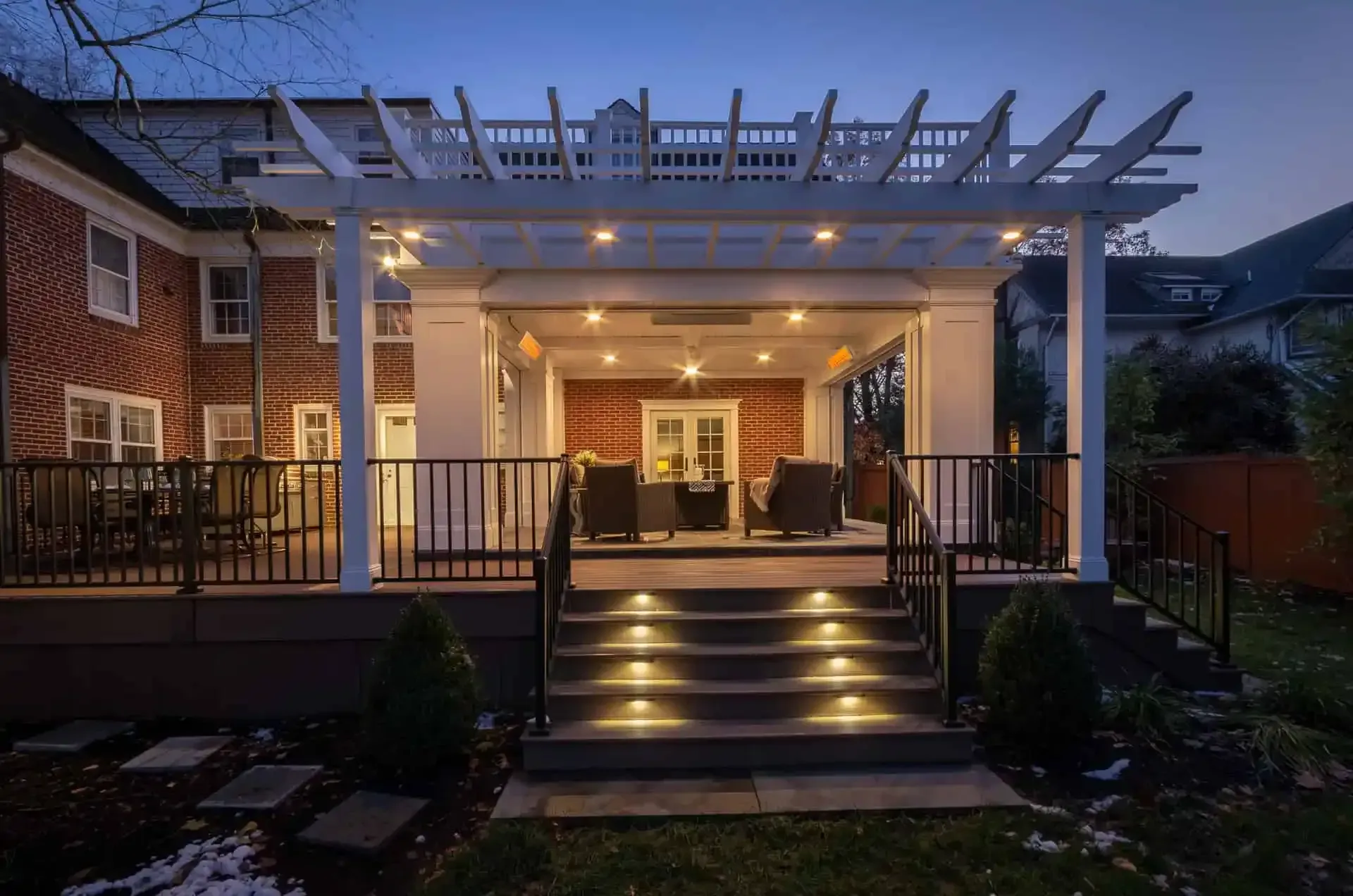 Backlit patio with pergola, steps, and outdoor seating beside a brick house at dusk