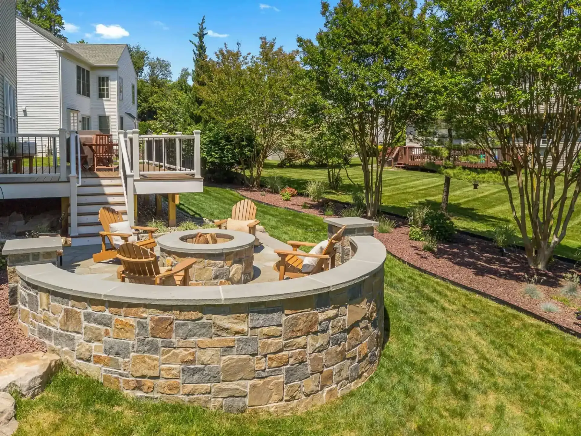 Backyard stone fire pit with curved seating wall beside a house and green lawn