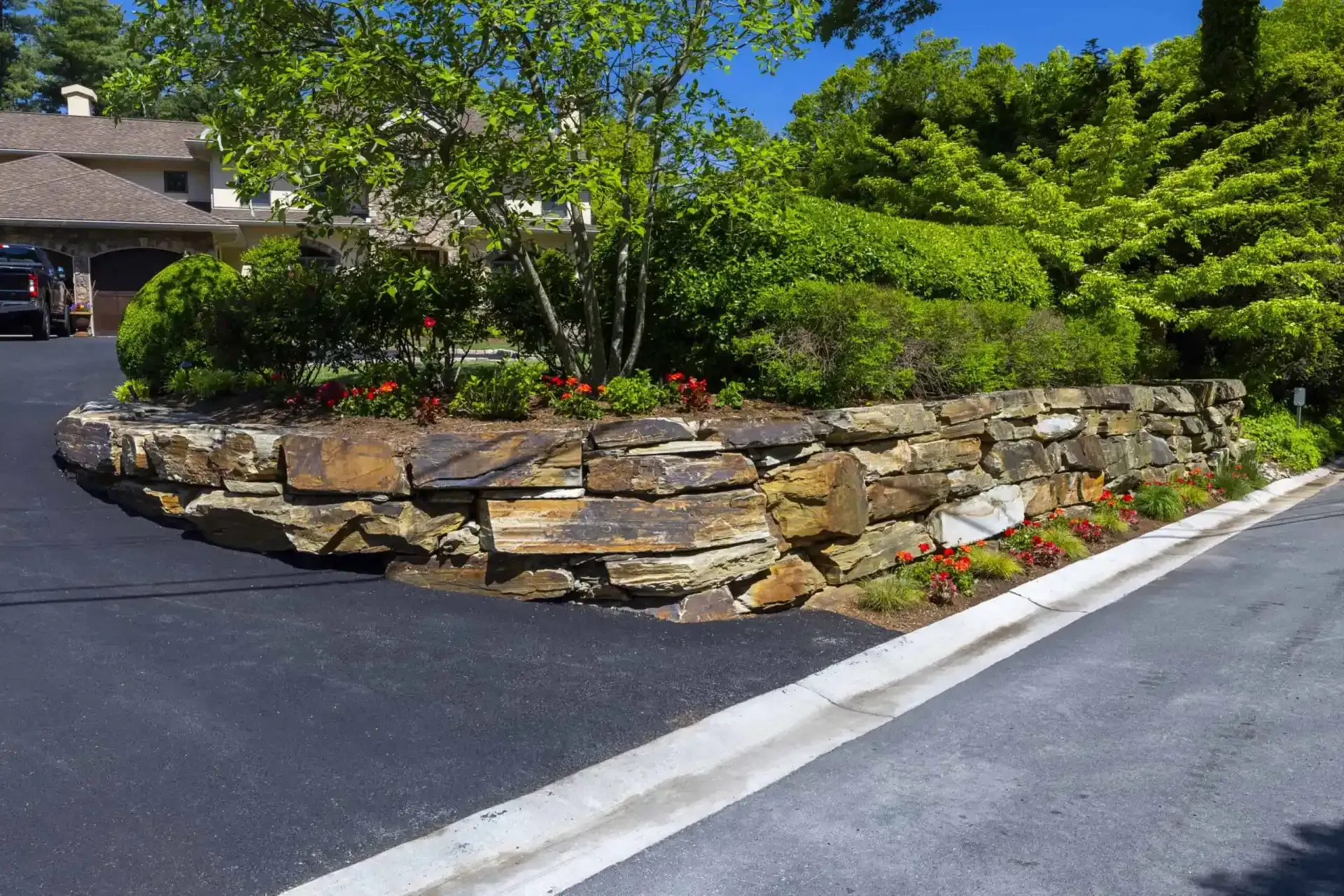 Stone retaining wall with shrubs and flowers beside a paved driveway in front of a building