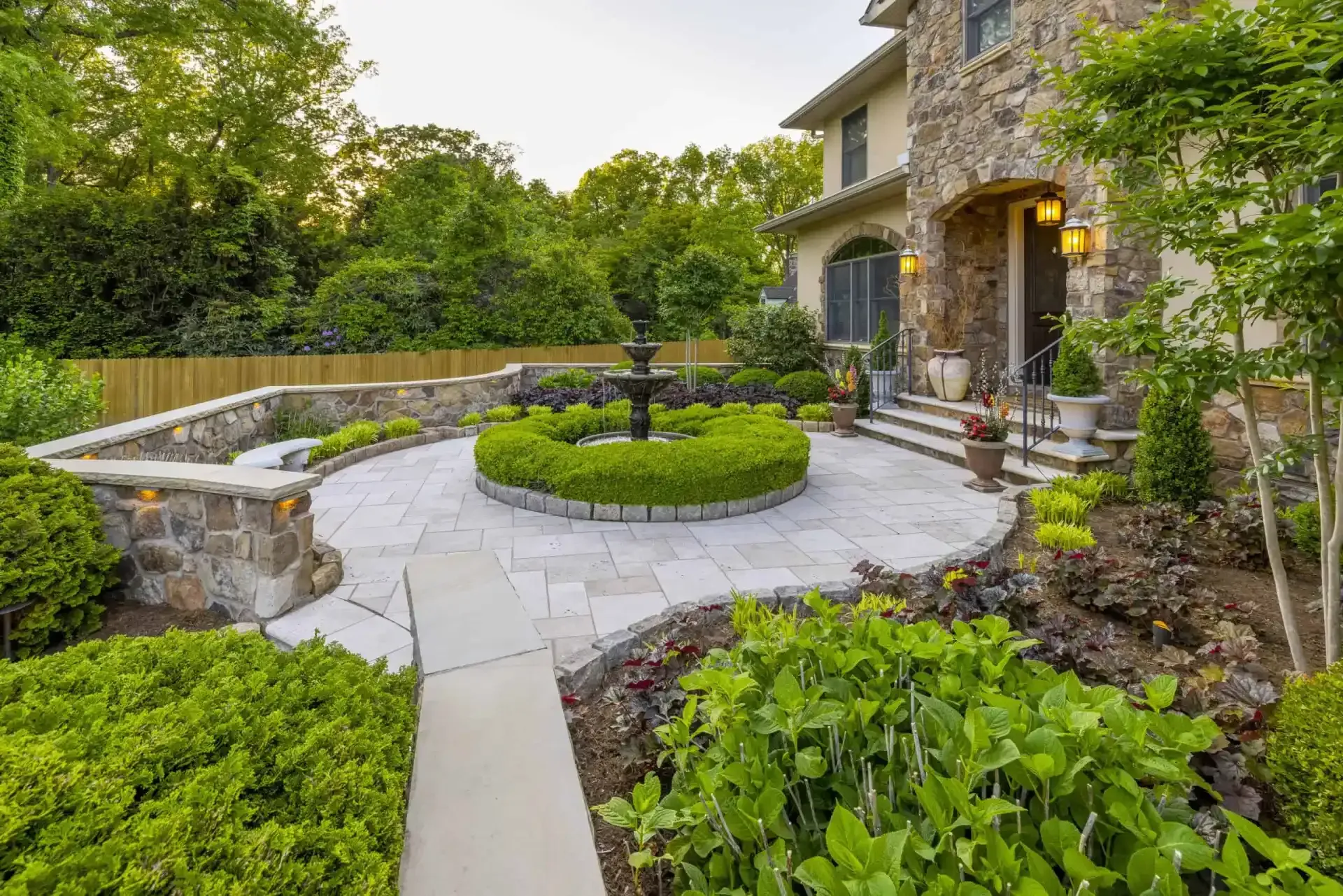 Stone patio and landscaped garden beside a house, with curved walkway, shrubs, and a fountain.