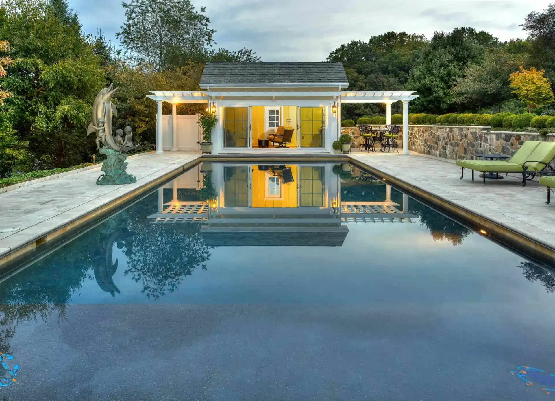Modern backyard pool with a reflective surface, stone deck, and a lit pavilion at sunset