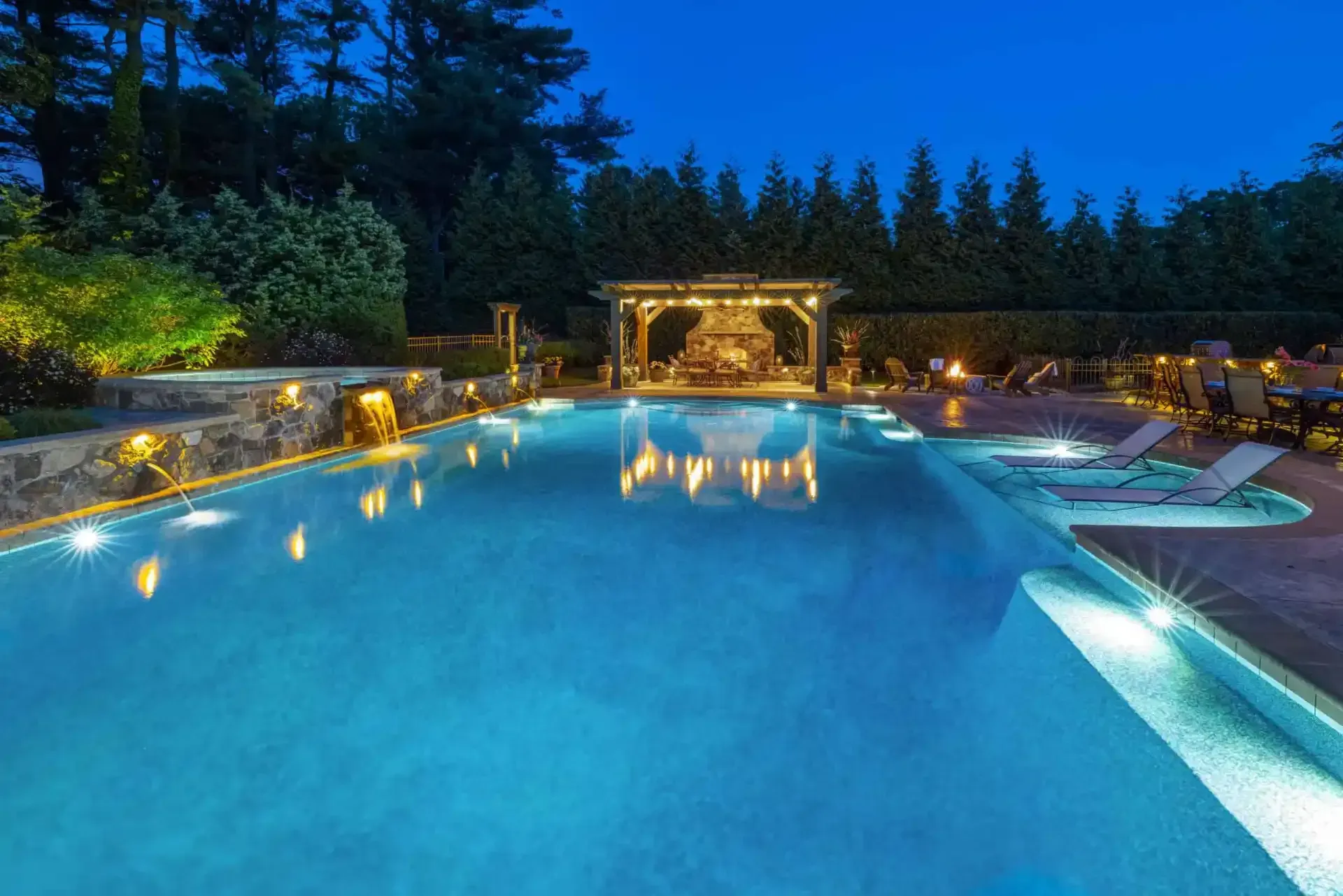 Illuminated outdoor swimming pool at dusk with lounge chairs and garden landscaping