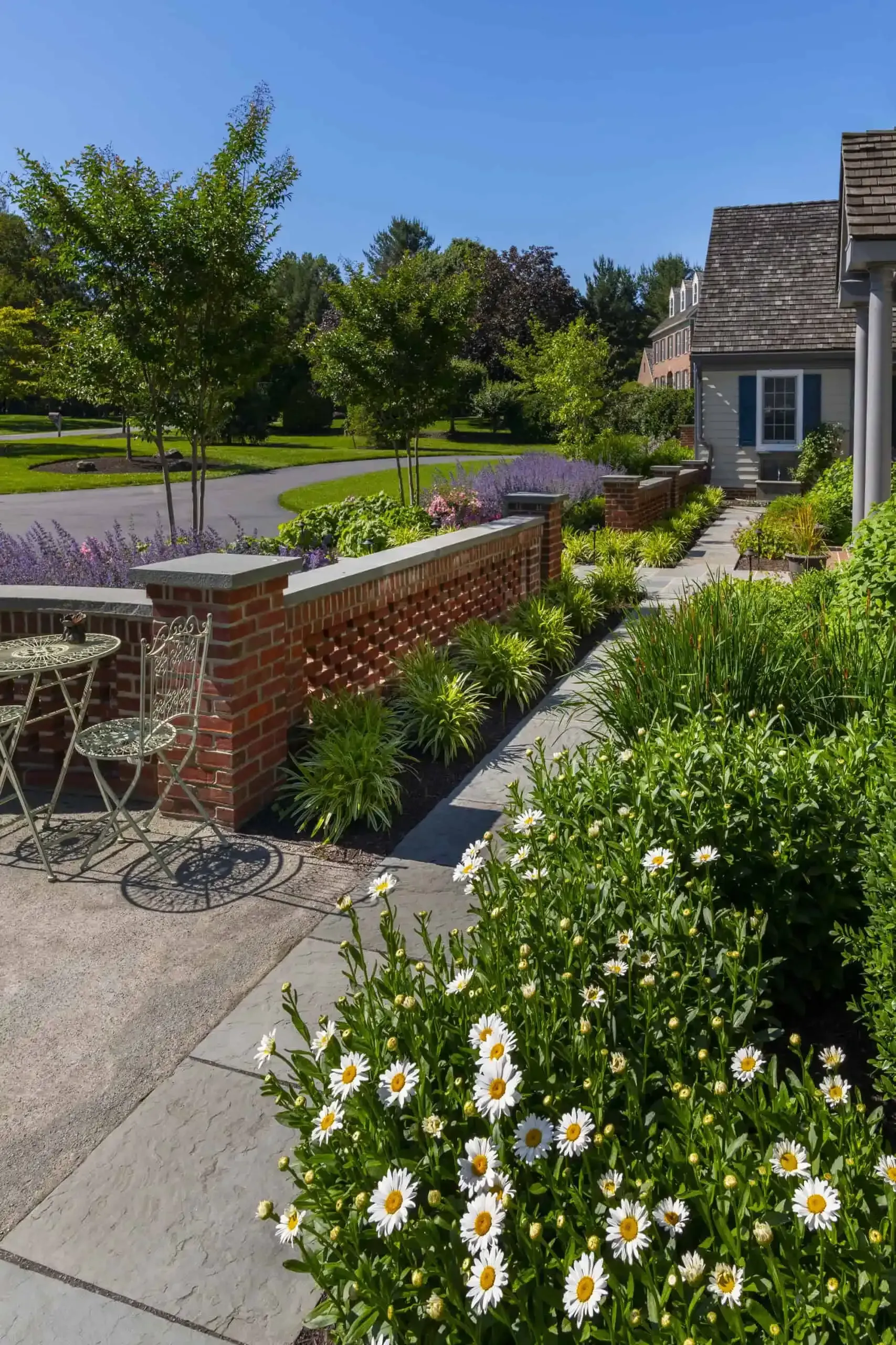 Garden patio with white flowers, brick wall, and a tree beside a lawn on a sunny day