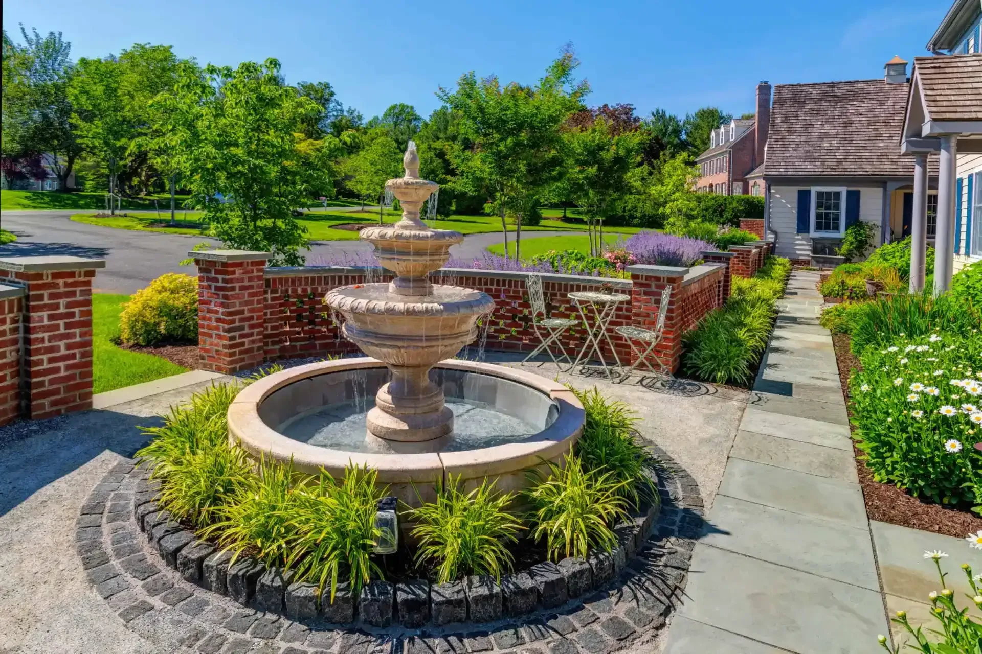 Tiered fountain in a brick courtyard with landscaped flower beds and suburban homes in the background