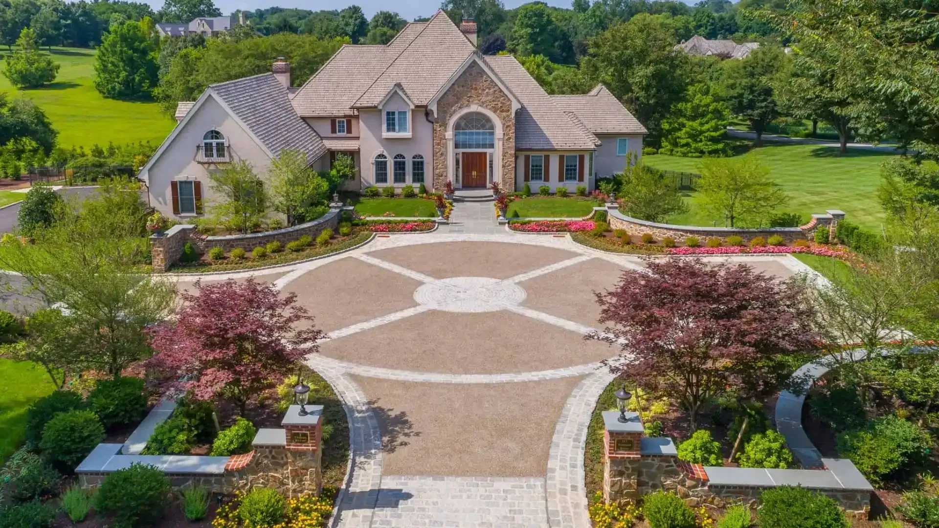 Large stone suburban house with circular brick driveway, landscaped garden, and red flowering trees in front yard