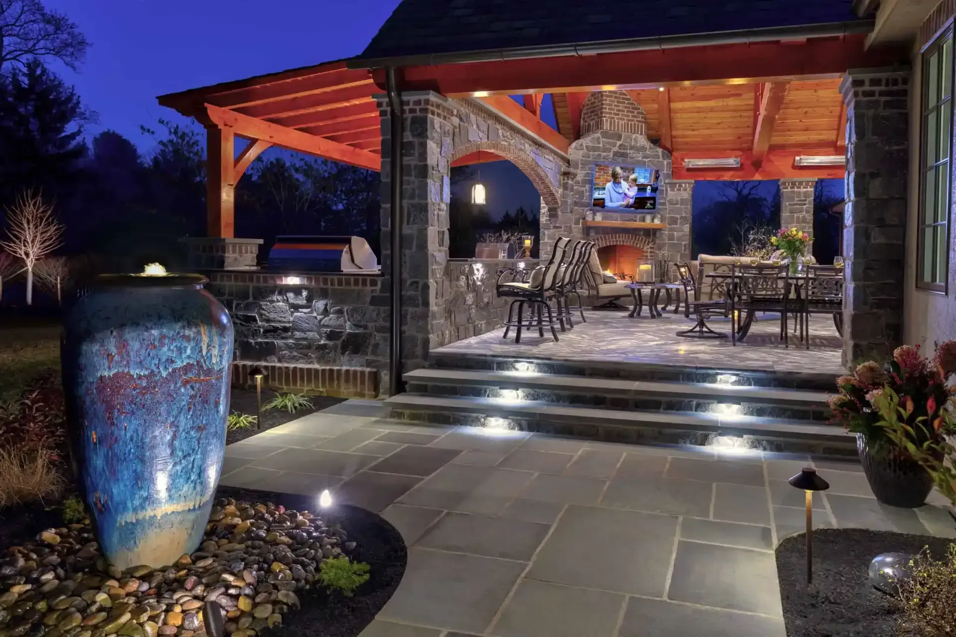 Covered patio at dusk with stone steps, outdoor tables, and glowing lanterns beside a large blue urn.