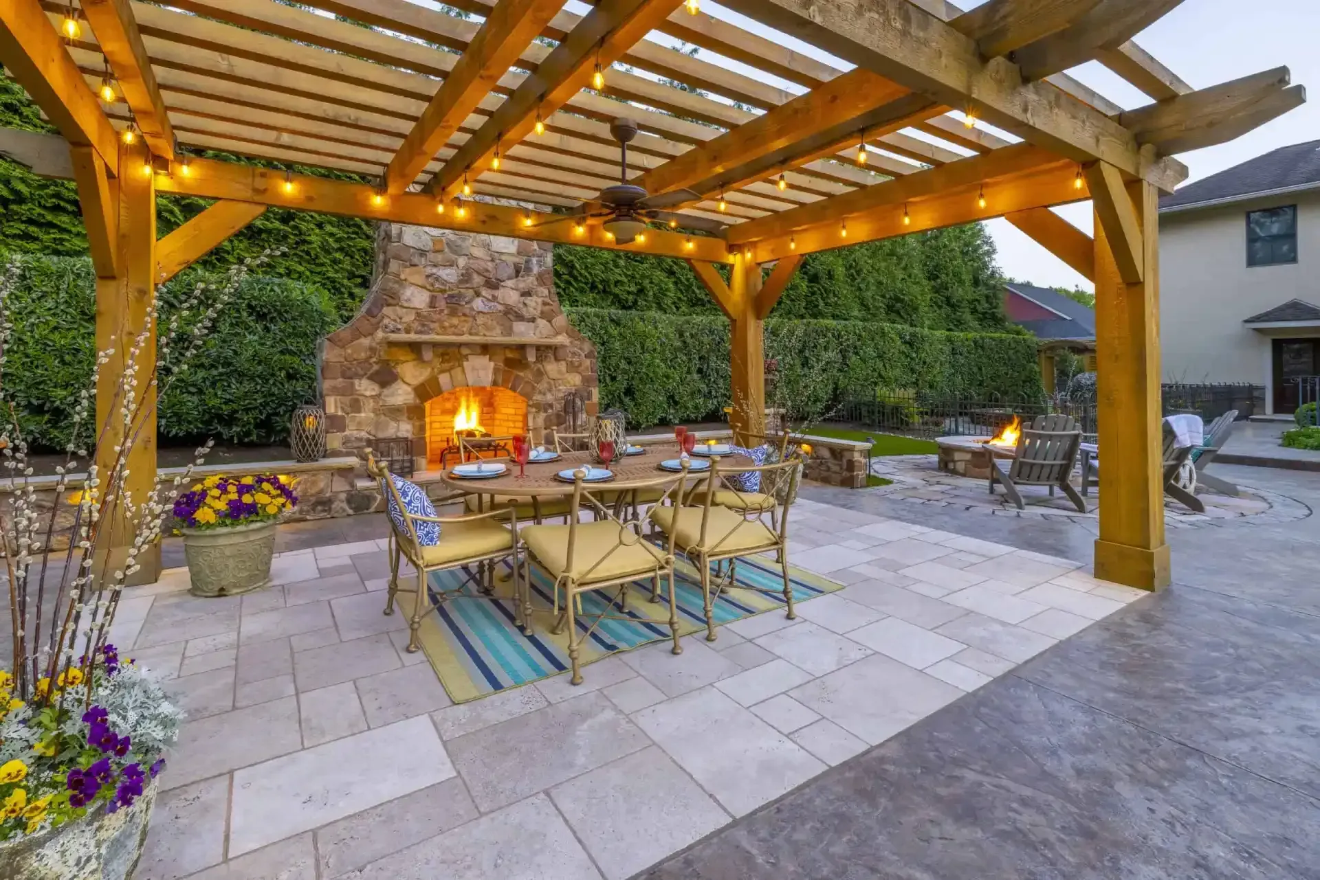 Outdoor stone patio with pergola, dining table, chairs, built-in fireplace, and colorful flower beds at dusk