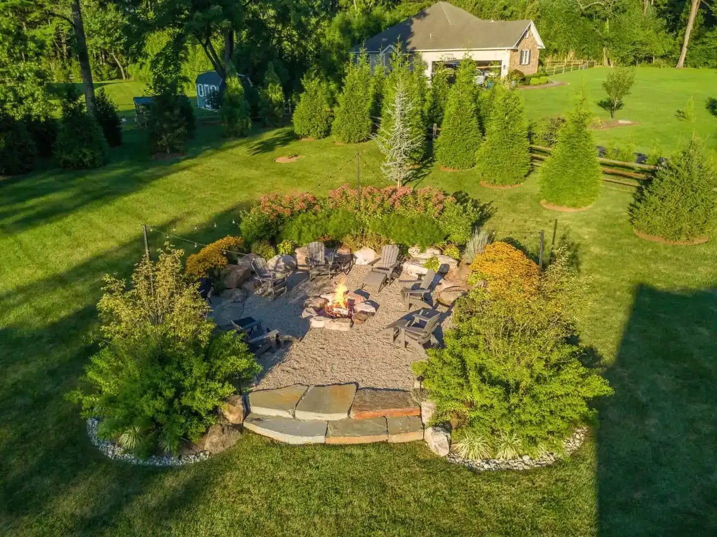 Backyard fire pit with stone seating area, surrounded by green lawn and trees, near a house