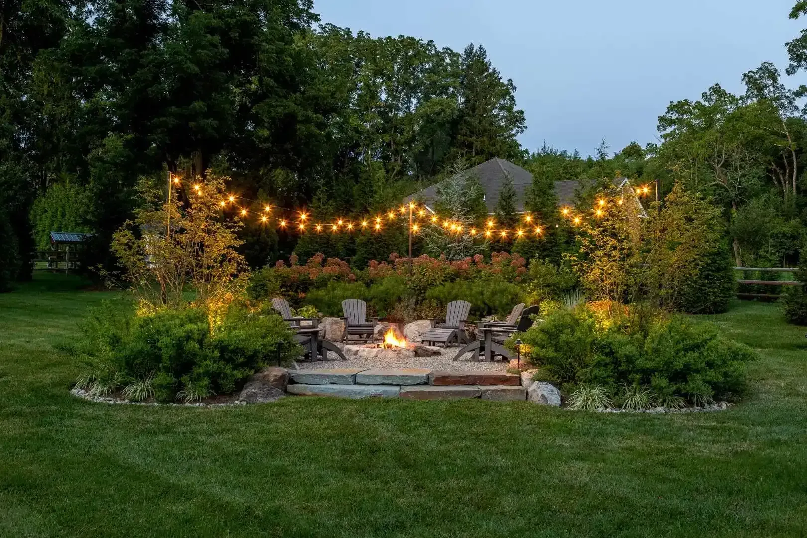 String lights over a backyard fire pit surrounded by chairs in a lush garden at dusk
