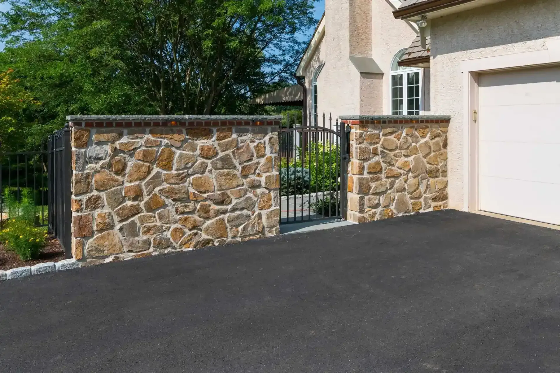 Stone wall and black metal gate beside a house garage driveway under bright sunlight