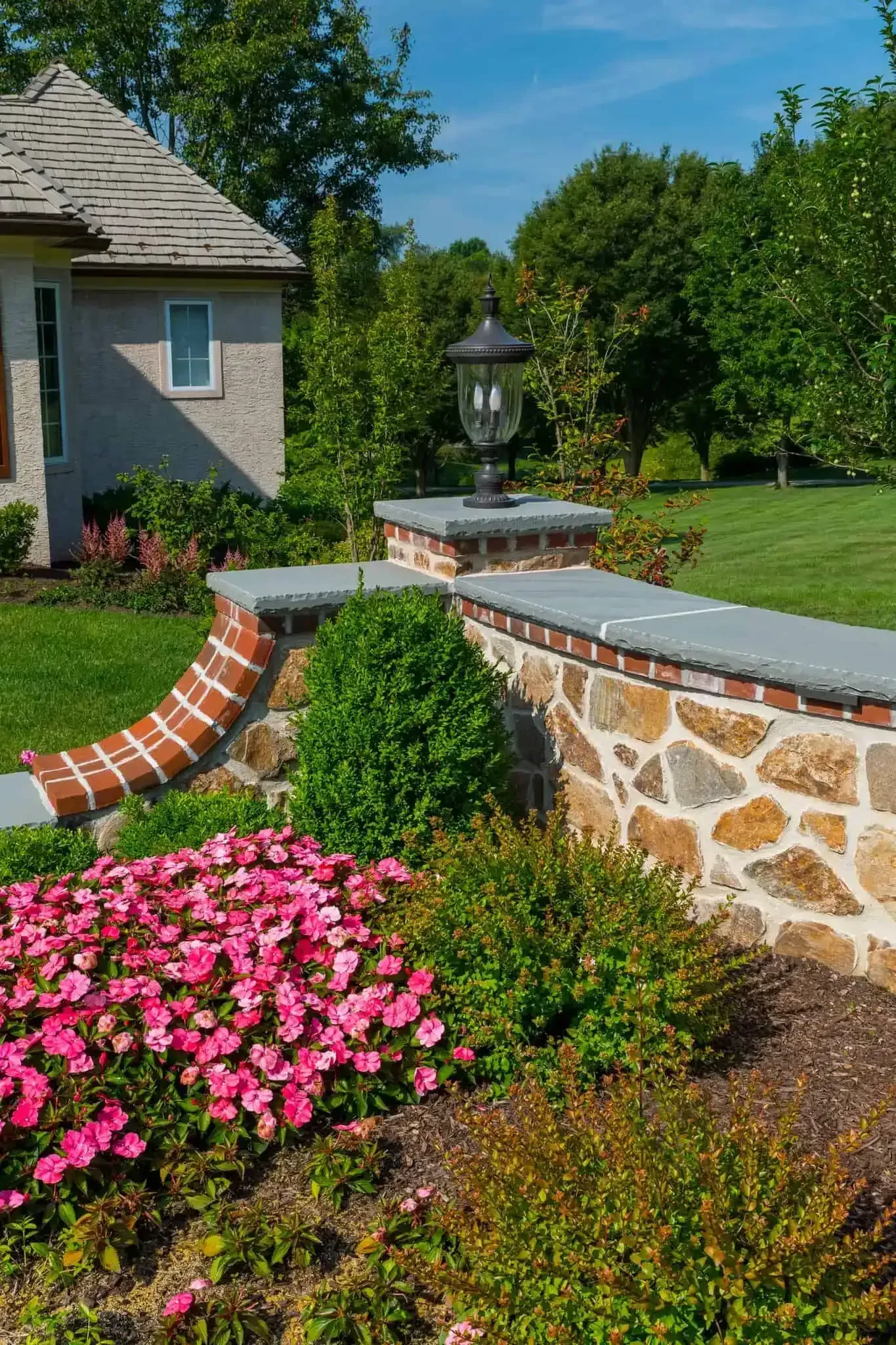 Landscaped front yard with stone wall, pink flowers, and a lantern by a house