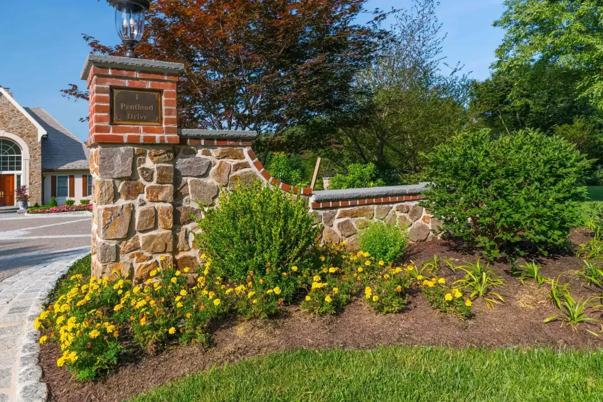 Stone entrance pillar with brick cap, surrounded by green shrubs and yellow flowers along a driveway