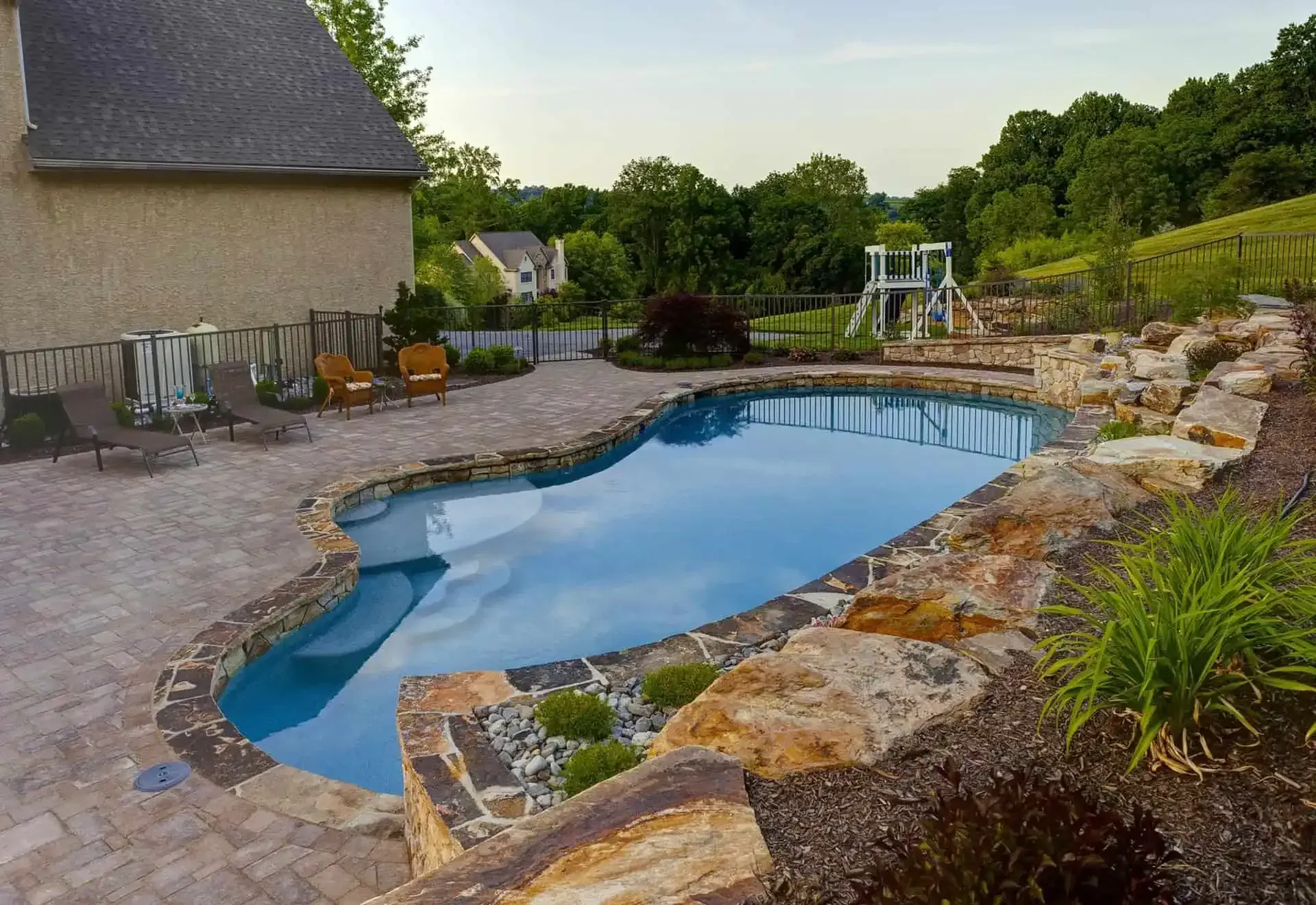 Backyard swimming pool with stone coping, patio chairs, and a swing set at sunset